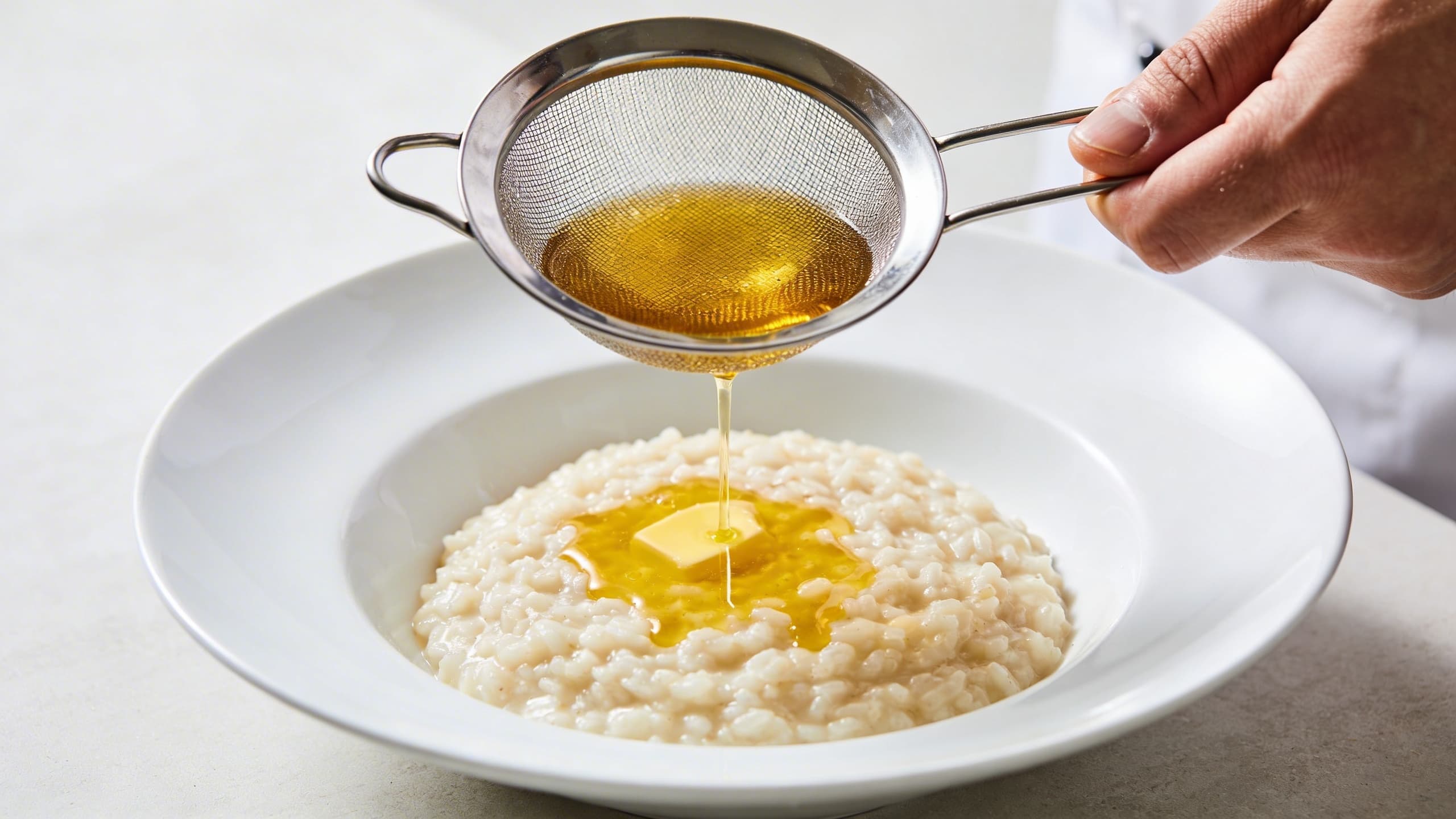 Image 2: A chef straining vibrant, galangal-infused golden butter through a fine-mesh sieve into a bowl of finished risotto.