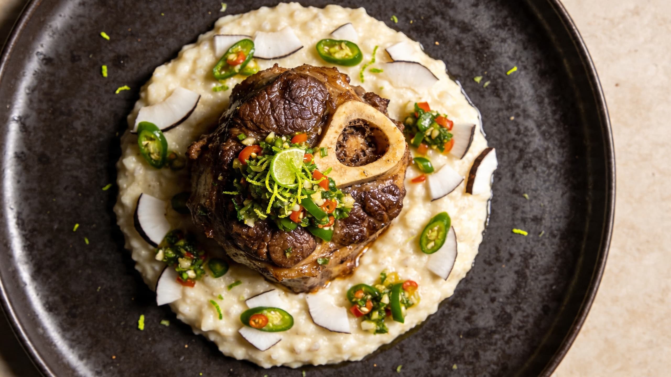 A beautifully plated Osso Buco shank resting on a bed of coconut-infused polenta, garnished with a bright green chili and lime gremolata