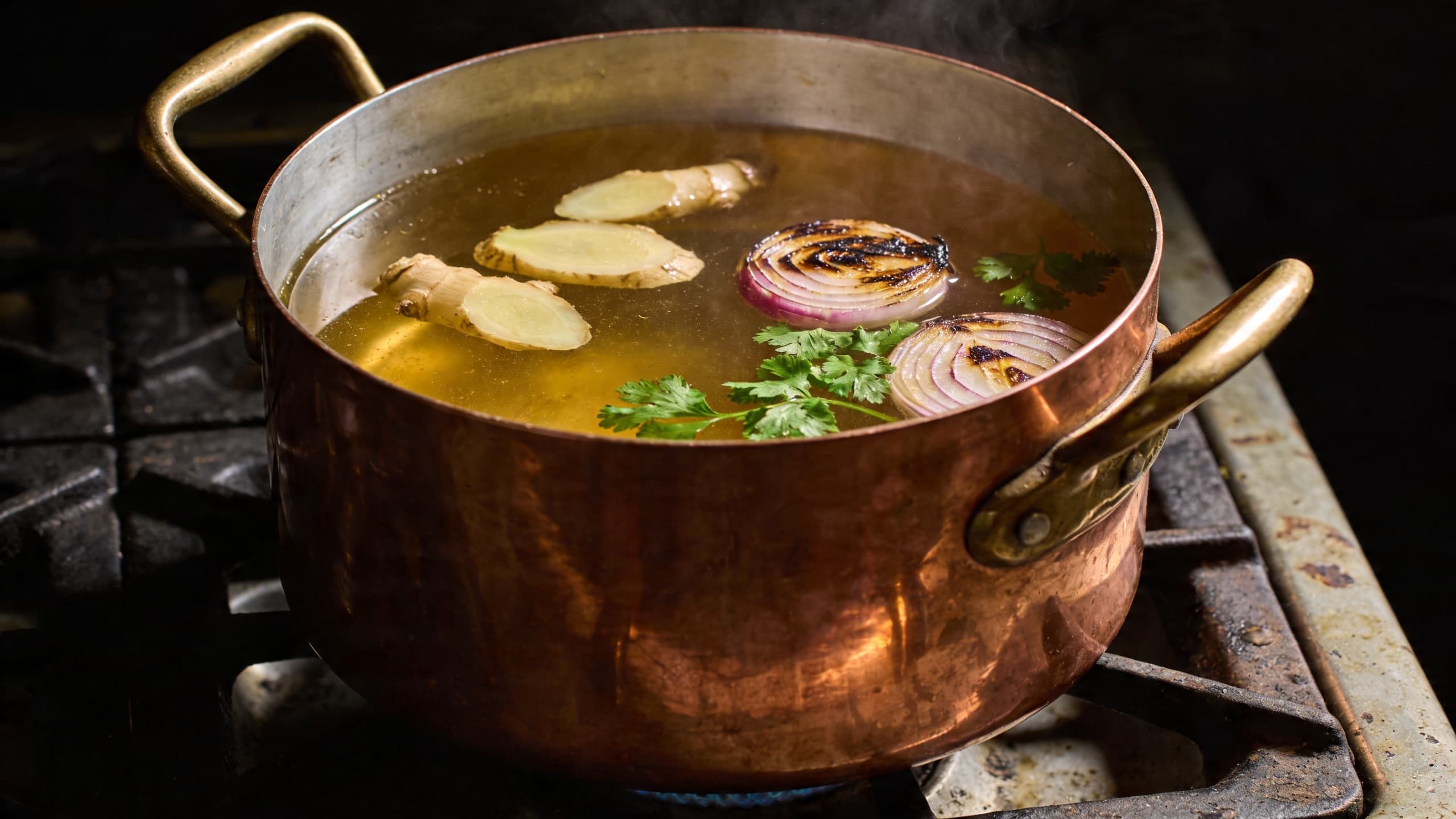 A large copper pot simmering a clear, golden broth with floating slices of galangal, charred onions, and fresh herbs.