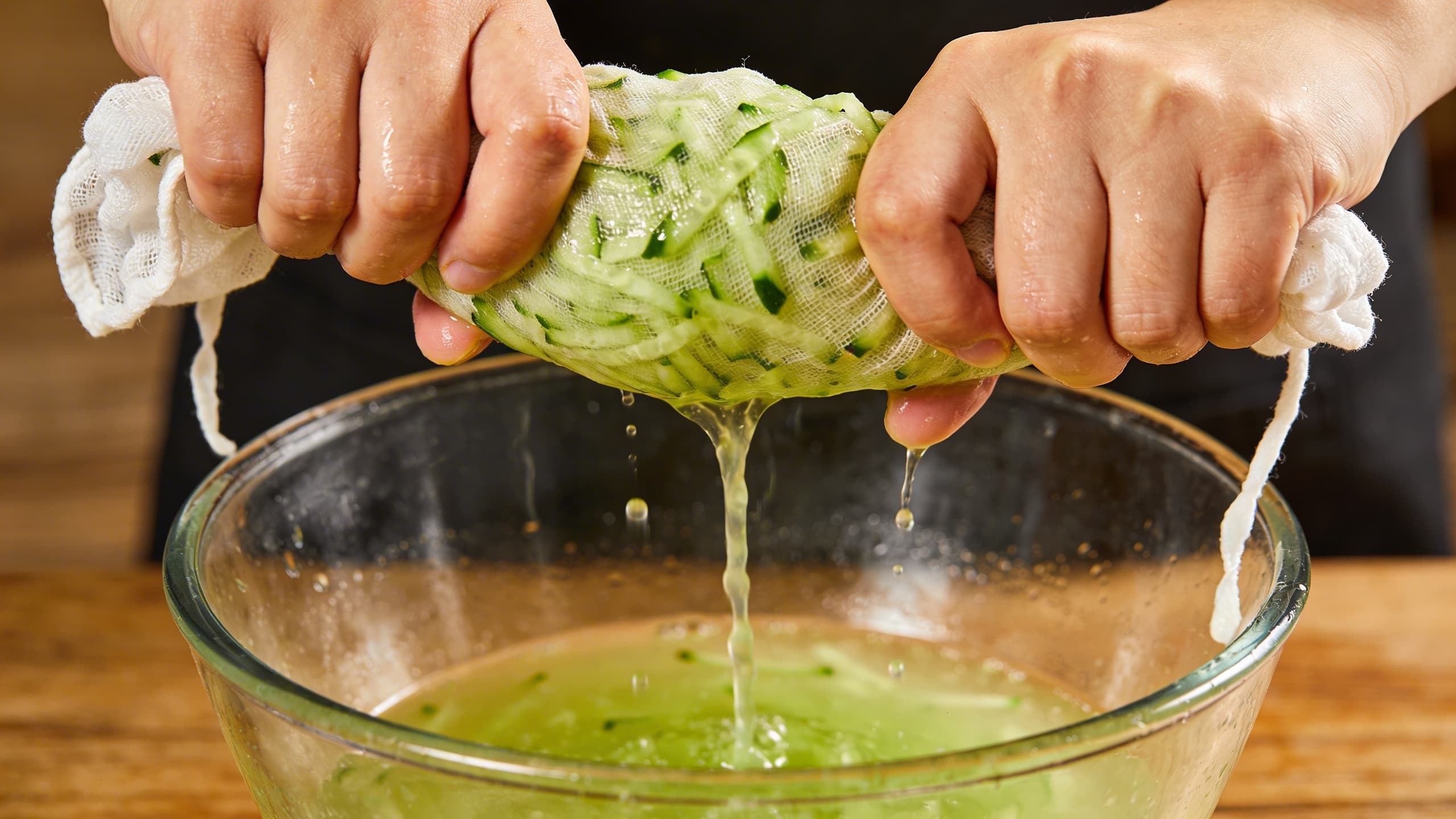 A close-up demo of squeezing grated cucumbers through a white linen cloth, showing the green juice being extracted.