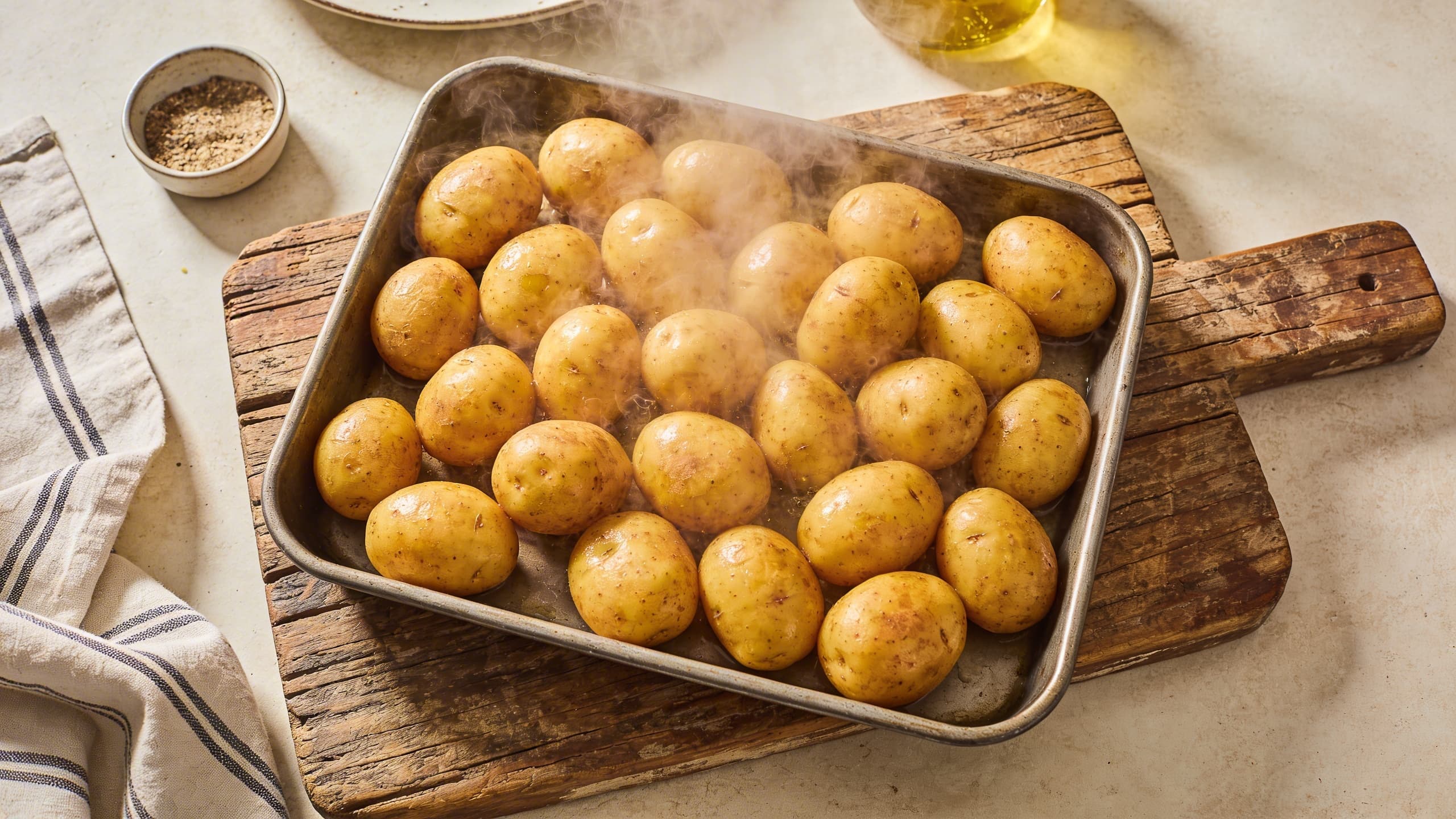 A tray of perfectly parboiled baby gold potatoes, steam rising, ready to be smashed on a rustic wooden board.