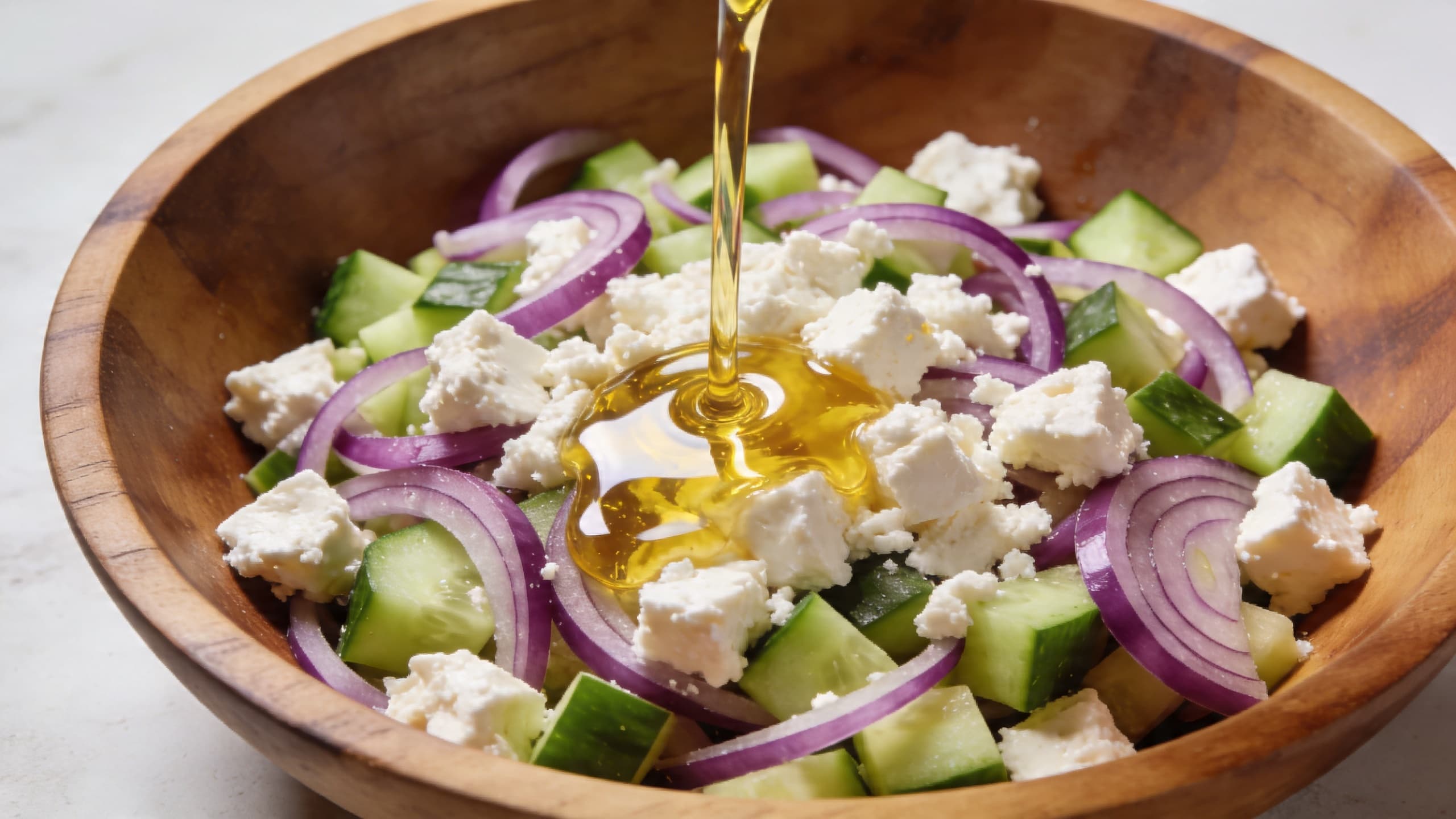Image 2: A close-up of golden olive oil being drizzled over a vibrant salad with feta cheese and red onions in a large wooden bowl.
