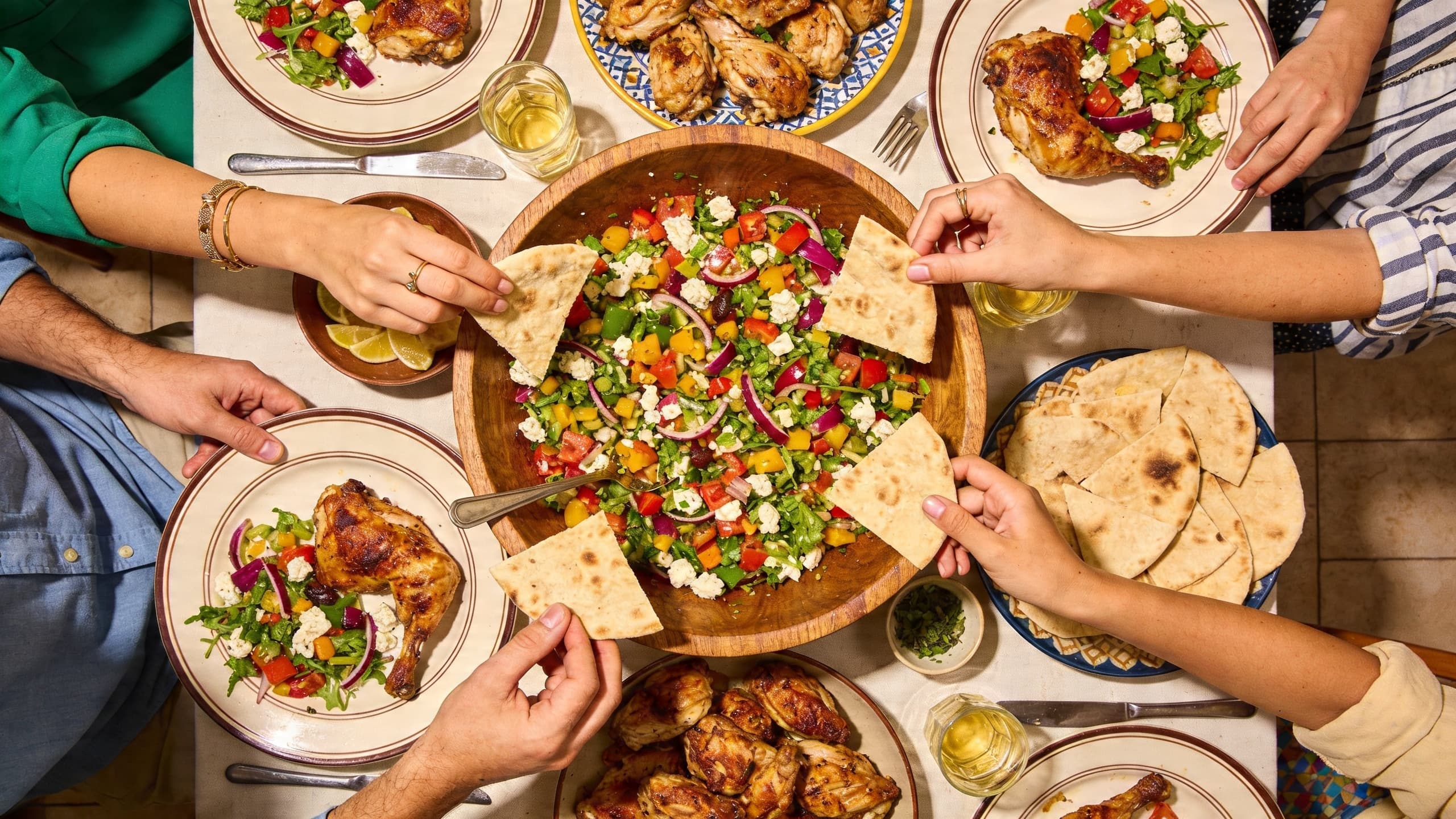 A vibrant family dinner table with a large bowl of Mediterranean salad and hands reaching for pita bread.