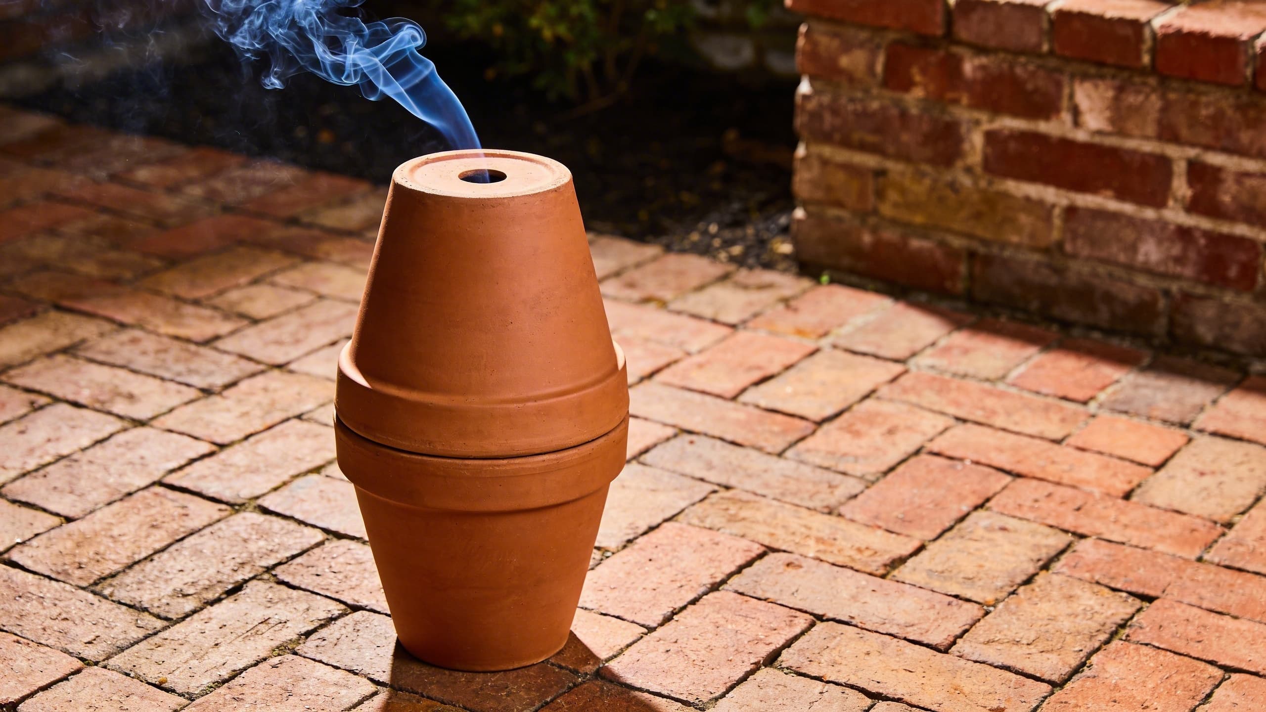 A DIY terracotta flower pot smoker setup with smoke gently curling from the top hole.