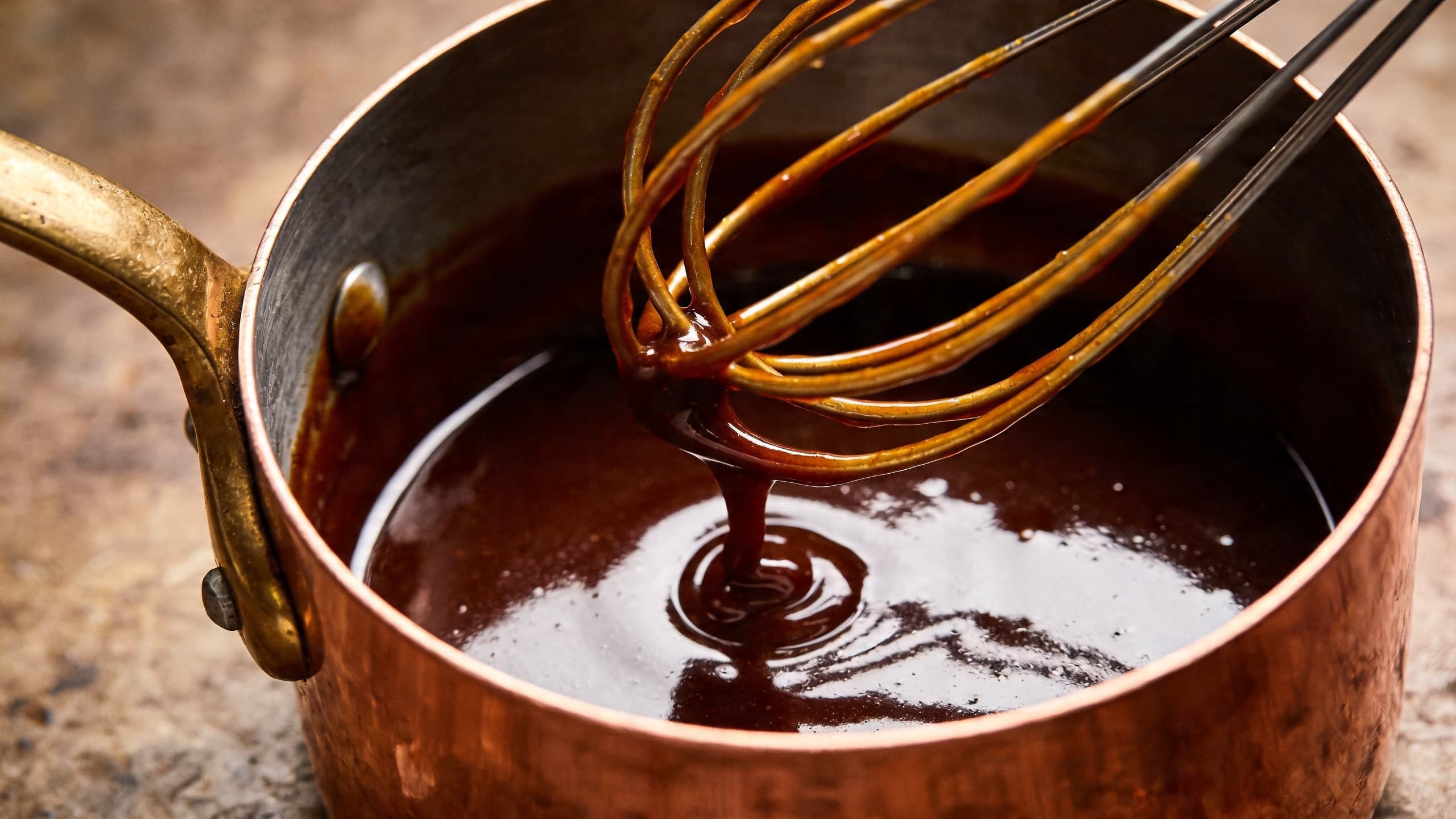 A glossy, deep mahogany smoked red wine reduction being whisked in a copper saucier, showing a perfect, mirror-like emulsion.