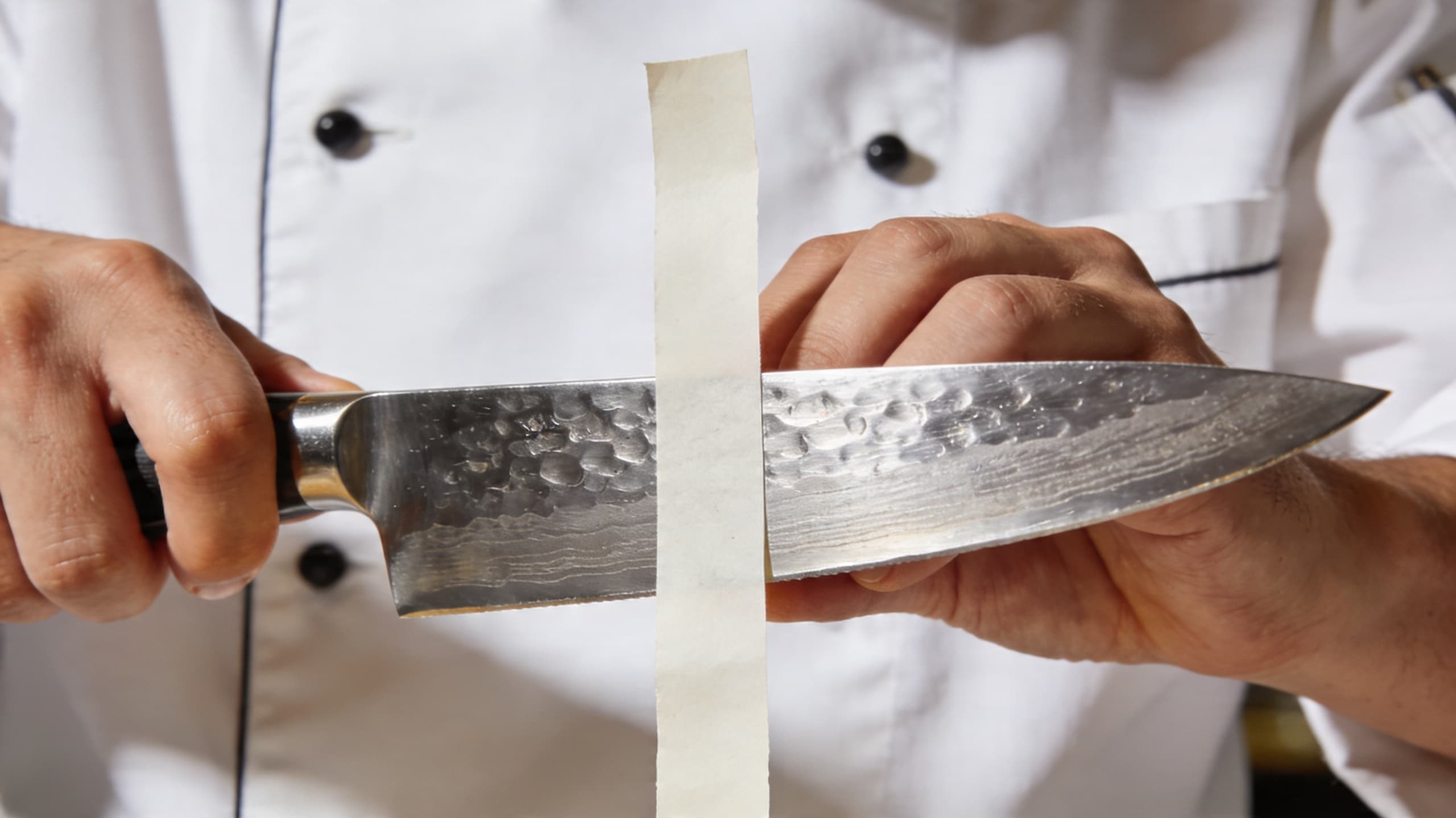 Image 2: A chef performing the 'paper test,' effortlessly slicing a thin strip from a vertical sheet of paper with a gleaming, polished chef's knife.