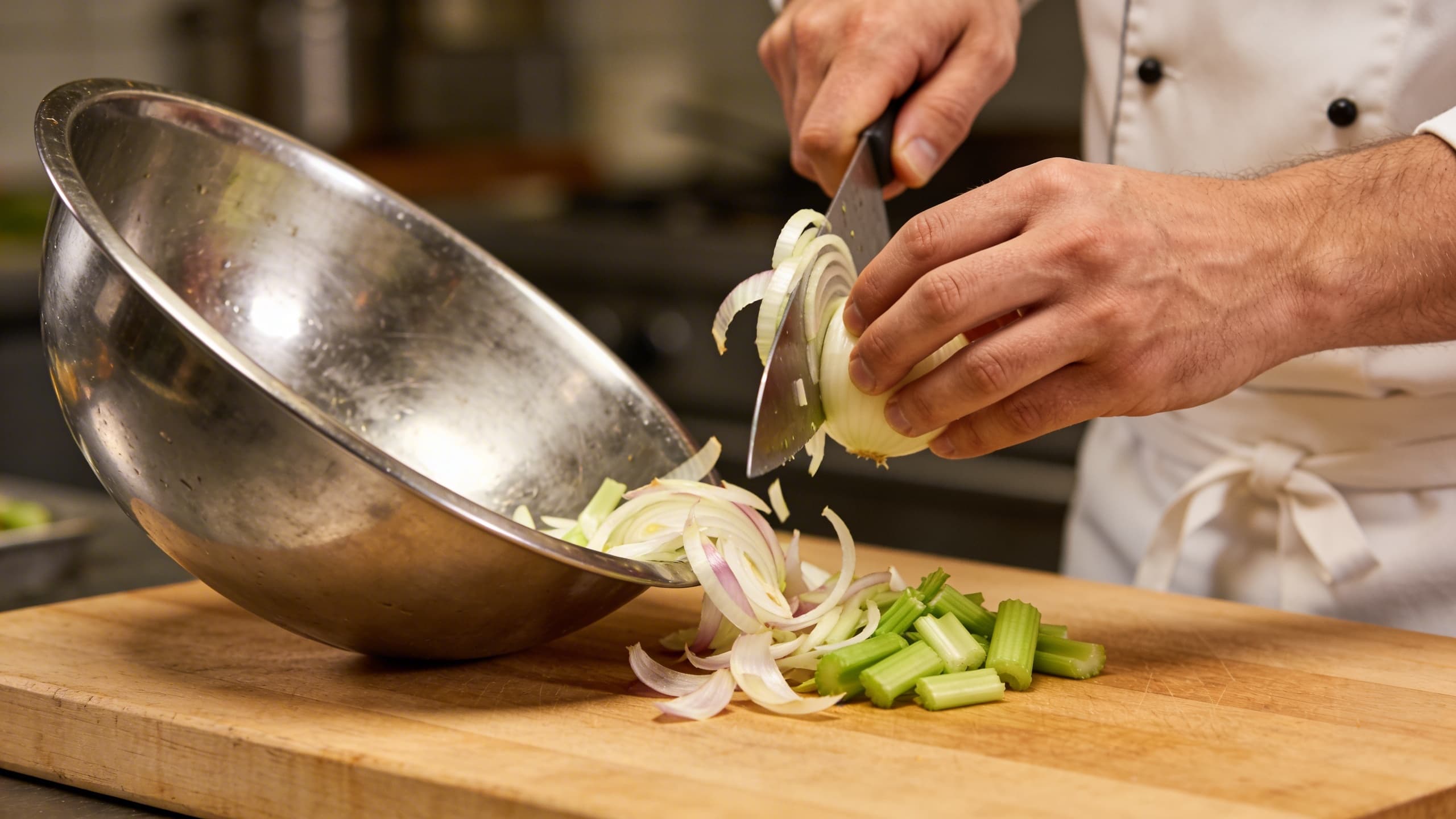 A chef's hand tossing onion skins and celery ends into a stainless steel scrap bowl next to a clean cutting board.