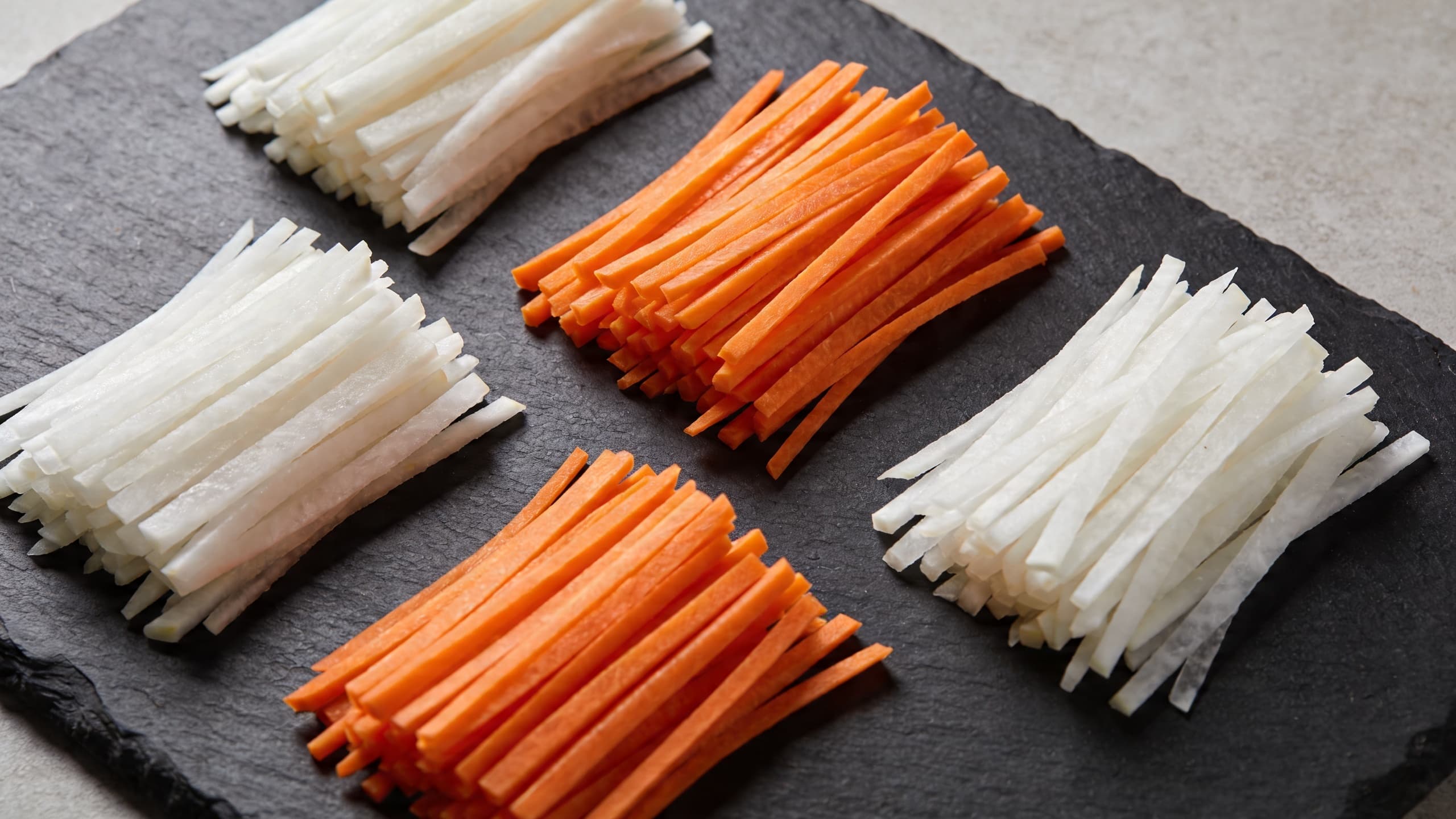 An overhead flat-lay of perfectly uniform julienned carrots and daikon radish on a dark slate board, showcasing the extreme precision of the matchstick cut.